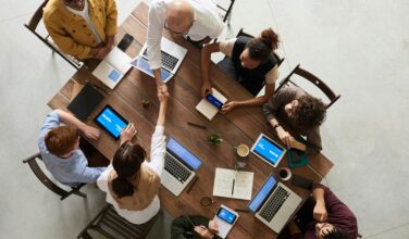 tafel van bovenaf gefotografeerd met 8 mensen, waarvan er twee elkaar de hand geven. Ondersteuning bij Juridisch Normenkader (JNK)