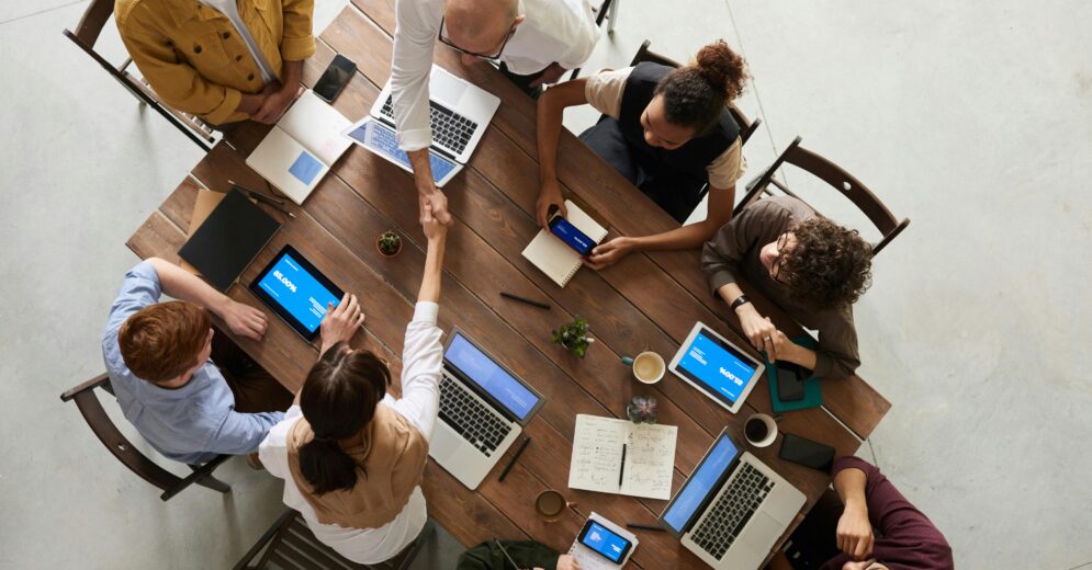 tafel van bovenaf gefotografeerd met 8 mensen, waarvan er twee elkaar de hand geven. Ondersteuning bij Juridisch Normenkader (JNK)
